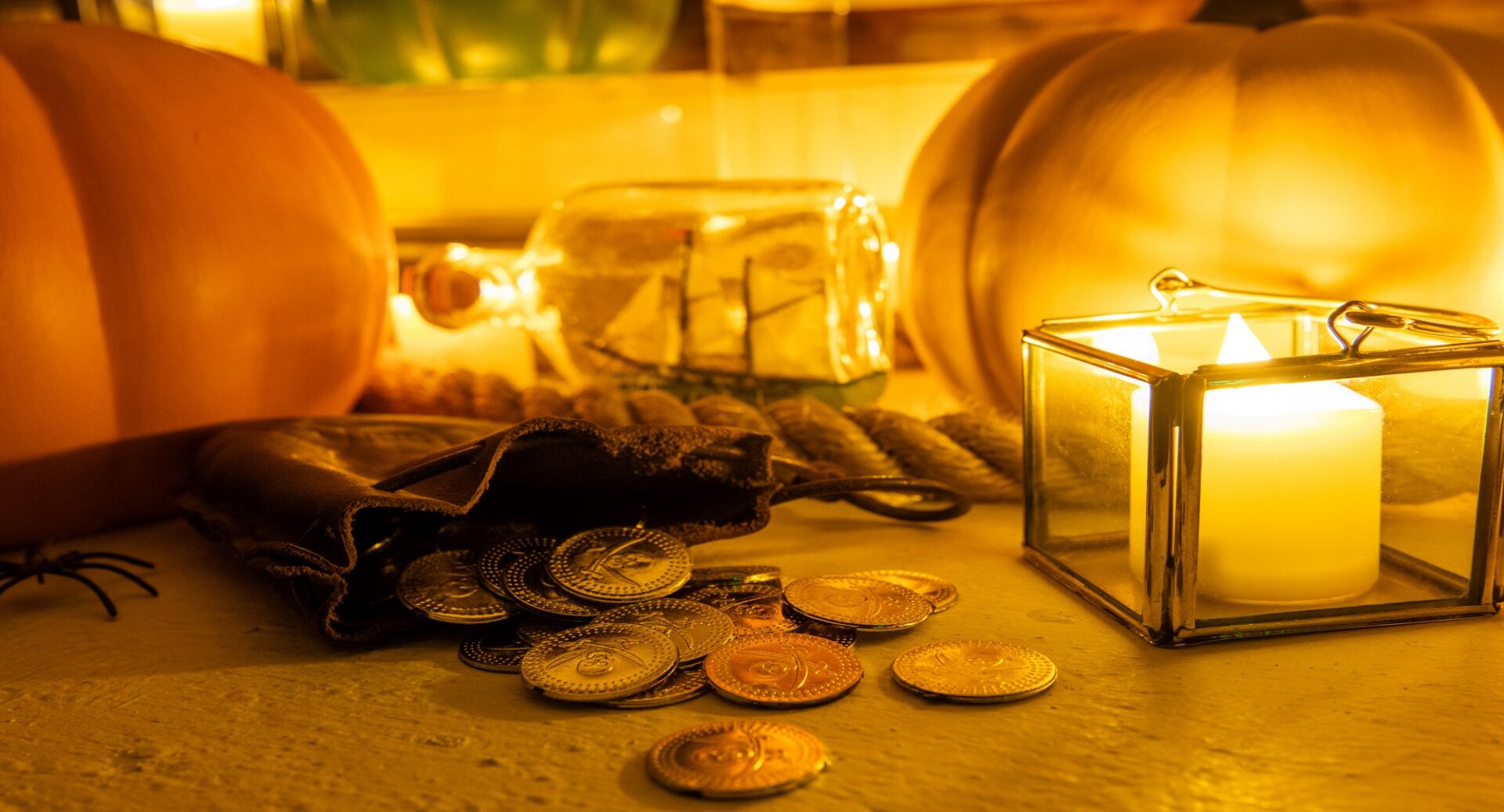 A candle-lit collection of pumpkins, gold coins and a ship in a bottle.