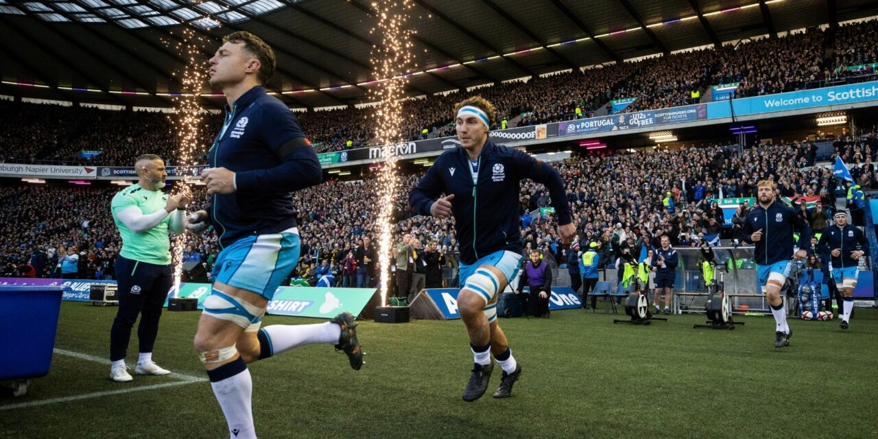 Rugby players running onto the pitch. Behind them can be seen a crowded stadium.