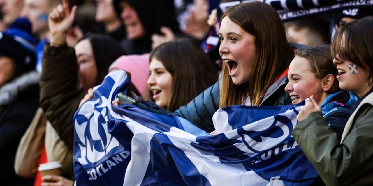 A crowd of people at a sports match, focusing on 4 girls holding a blue and white flag.