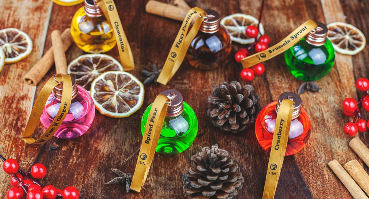 Variety of glass baubles filled with gin, fir cones, red berries and cinnamon sticks on a wooden worktop.