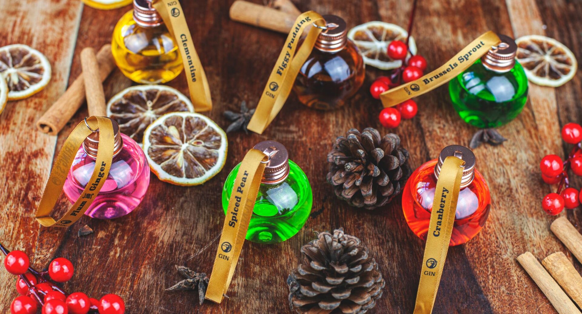 Variety of glass baubles filled with gin, fir cones, red berries and cinnamon sticks on a wooden worktop.