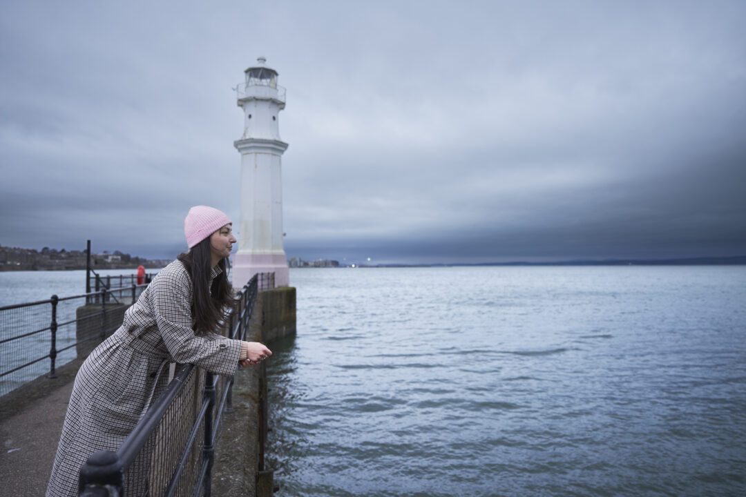 Woman leans on the barrier at Newhaven lighthouse on a wintery day, looking out across the harbour.
