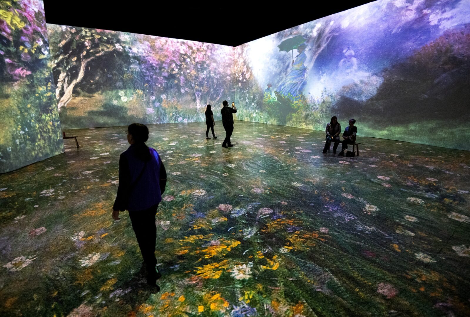 People standing in a darkened room, in which the walls show projections of a painting filled with flowers.