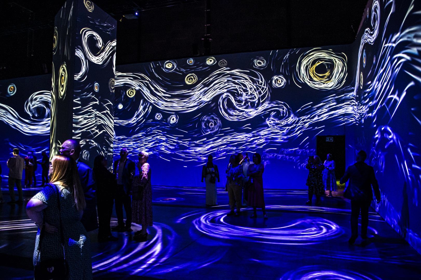 People standing in darkened room, the walls of which have projections of white swirls on a dark blue background