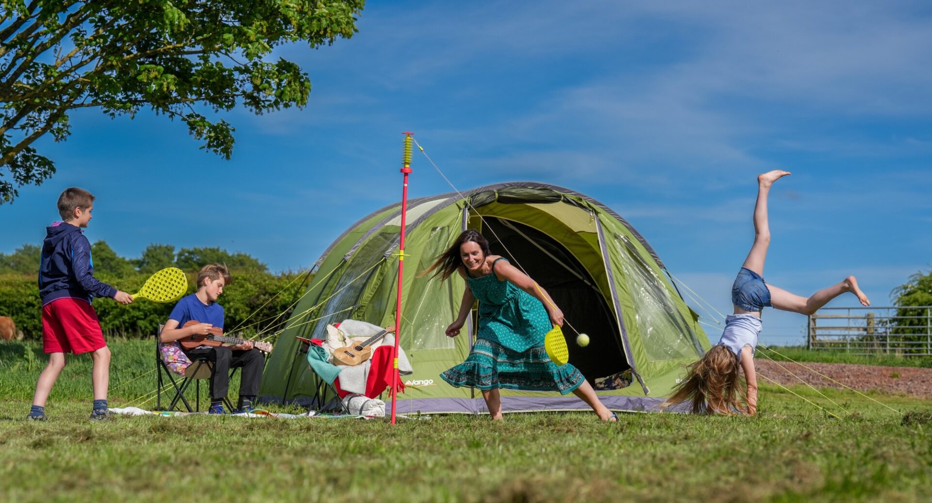 Lady playing swingball with son outside her tent while daughter is doing a cartwheel and other son is playing guitar while sitting on a camping chair.
