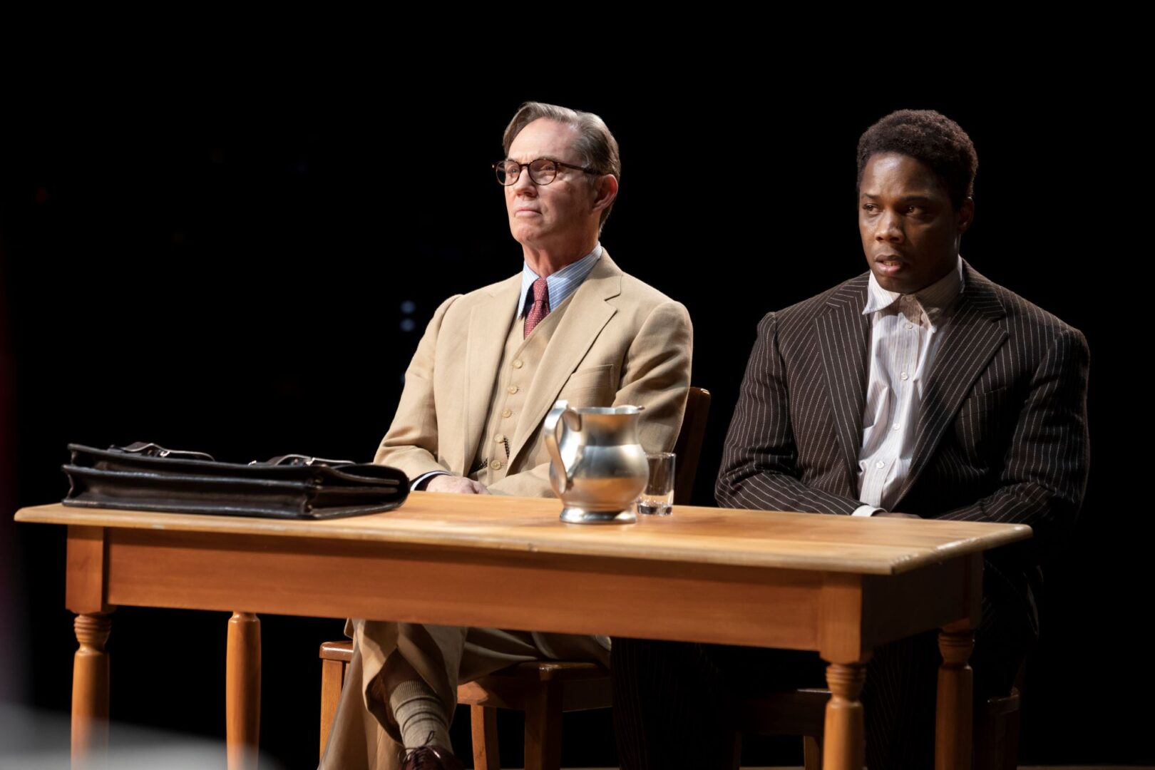 2 men sitting behind a desk, on which there is a briefcase, a jug and a glass of water.