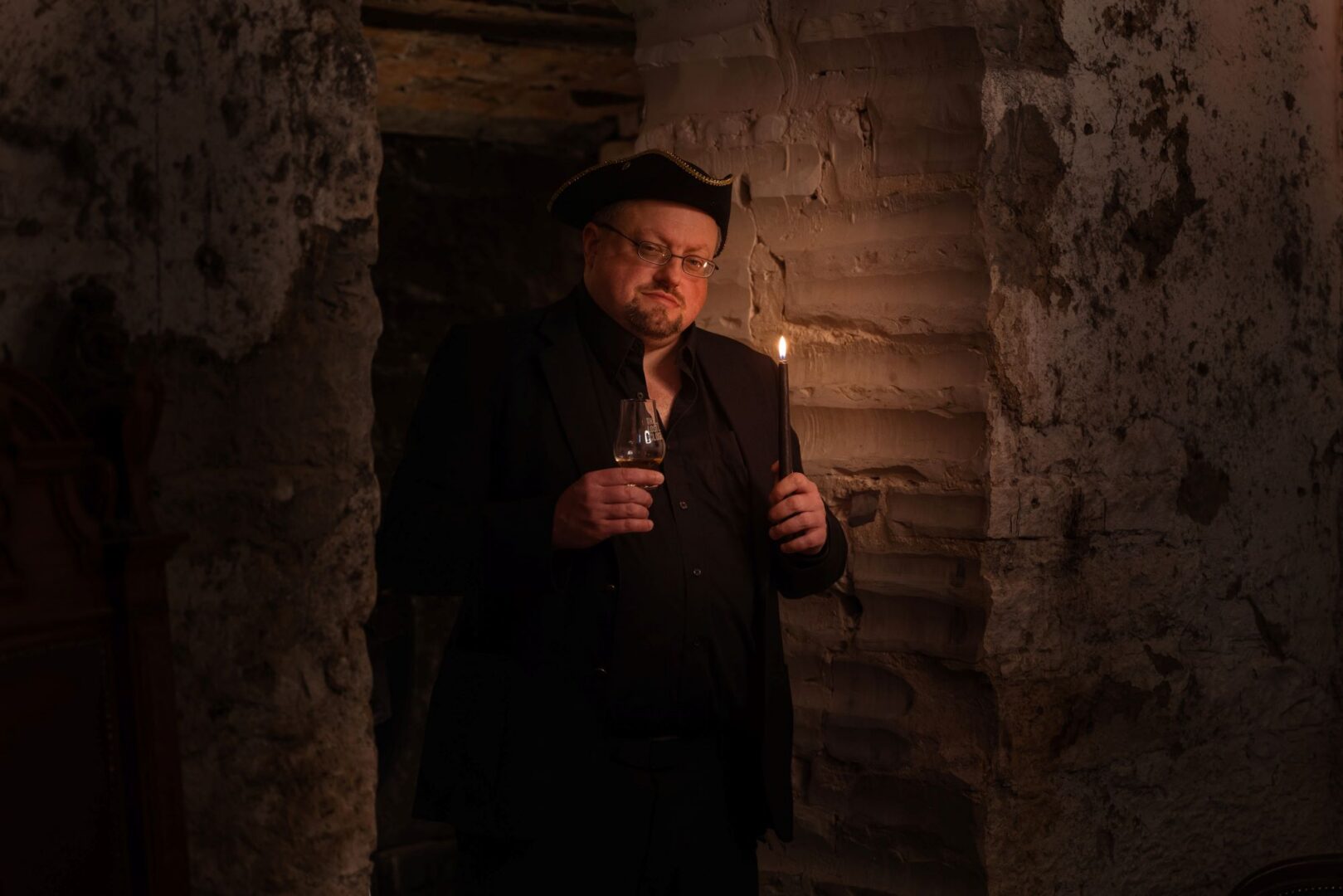 Man standing in an underground dark space, holding a glass of whisky and a lit candle.