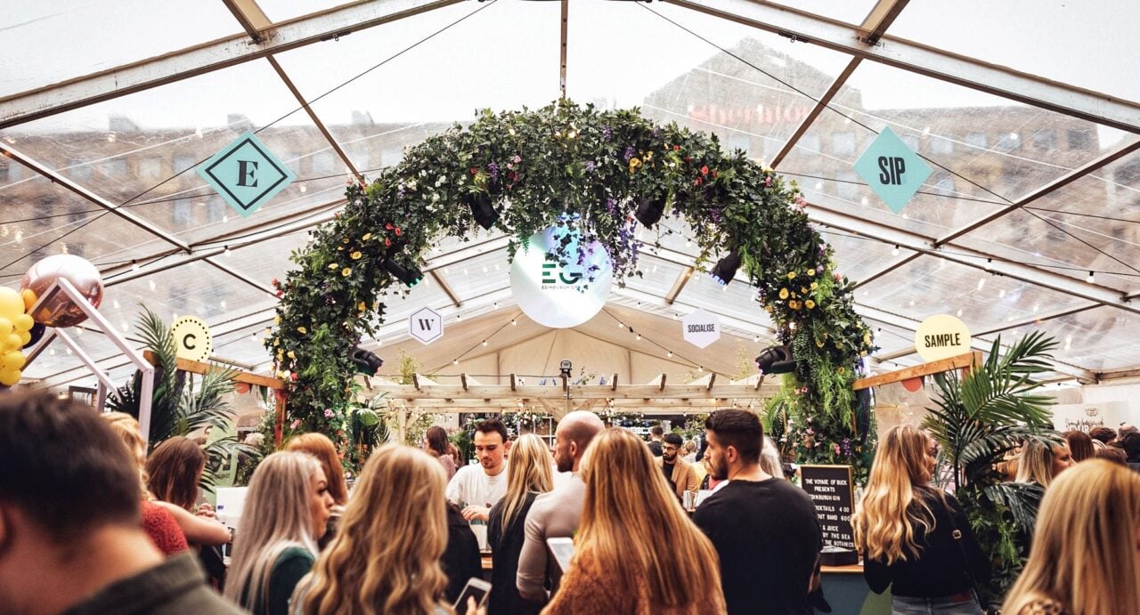 Crowd of people standing in front of a bar, as a barman serves drinks. Overhead, is a glass ceiling, and a garland of flowers over the bar.