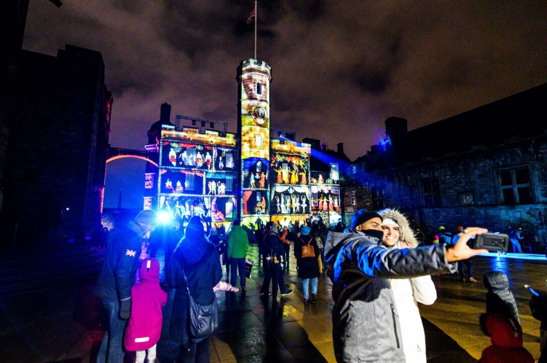 Groups of people standing outside, against the backdrop of a building covered in illuminations.