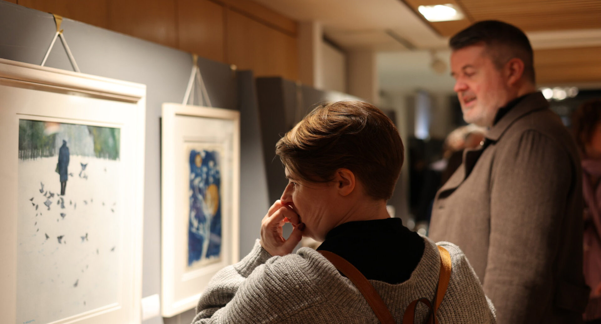 2 people looking at a row of paintings hanging on a wall.