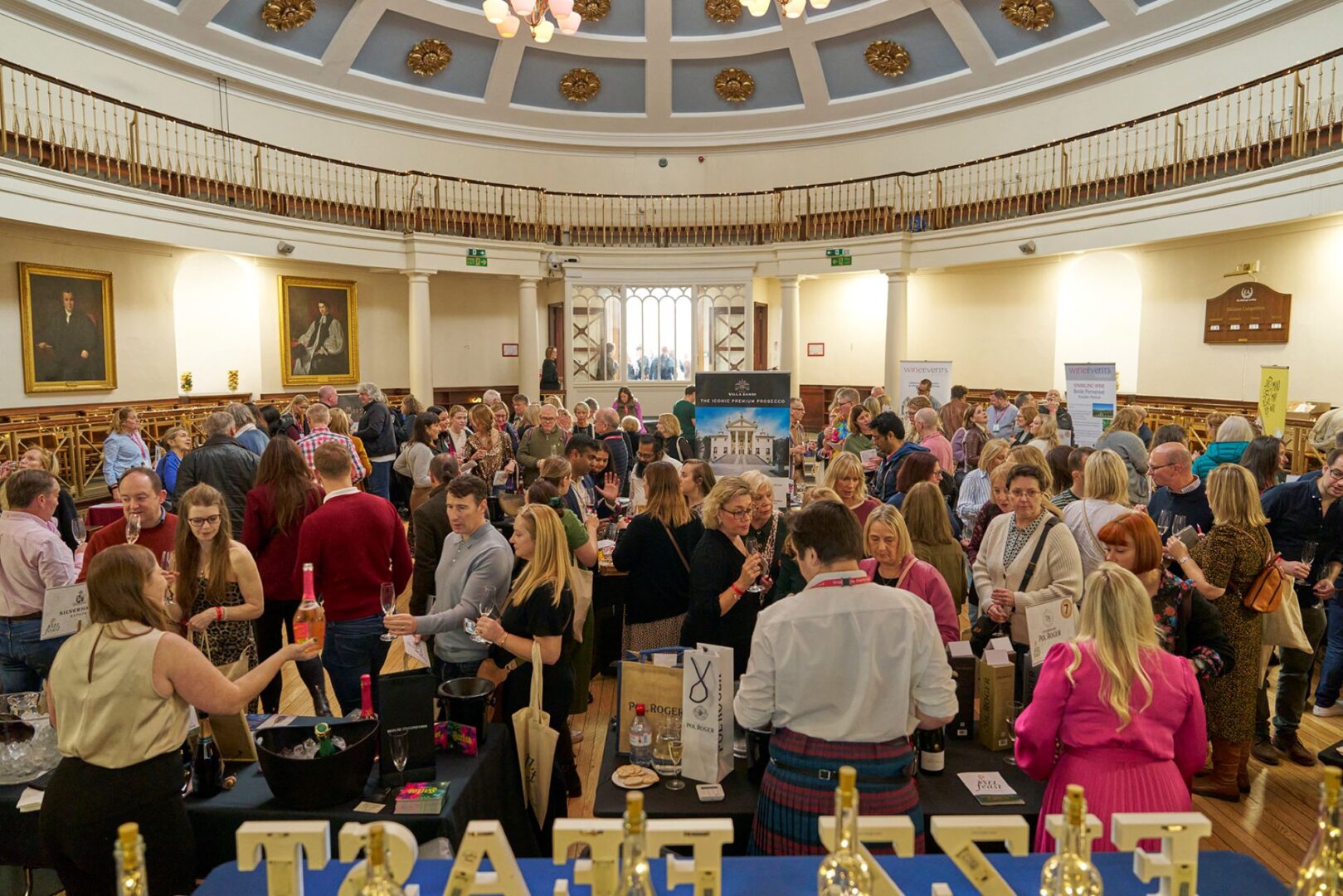 A crowded room of people wandering round tables and tasting drinks.