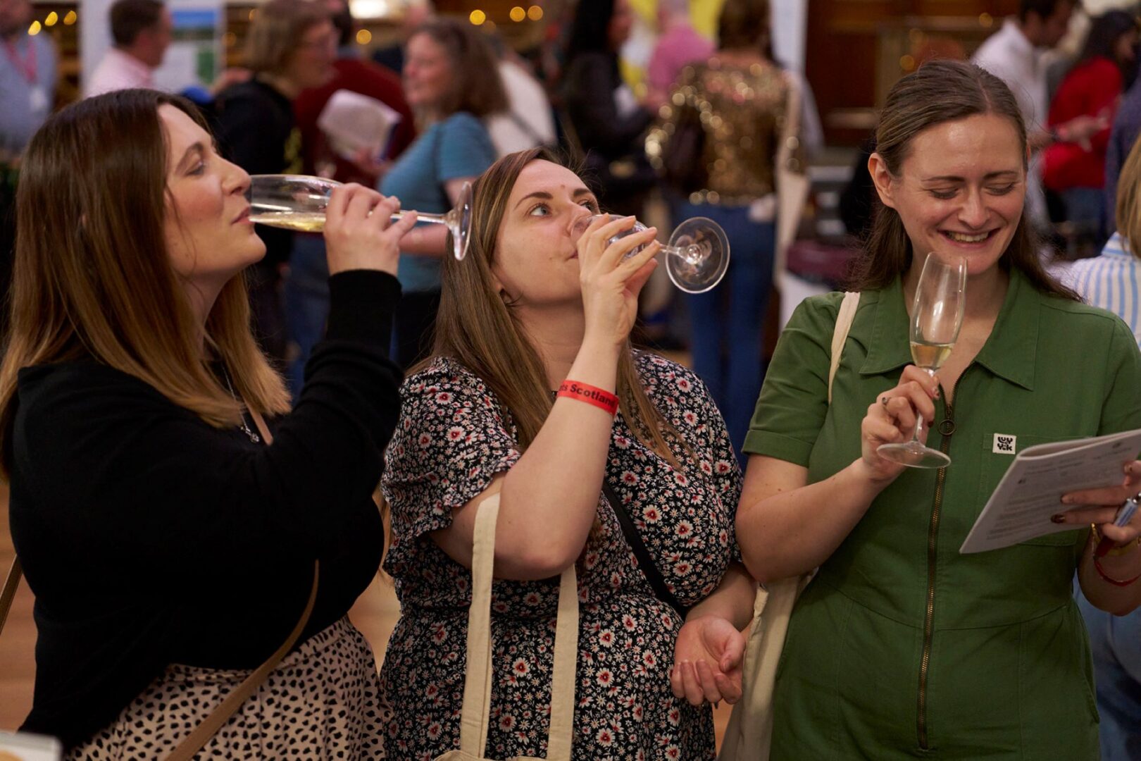 3 women standing in a crowded rooms, sipping from wine glasses.