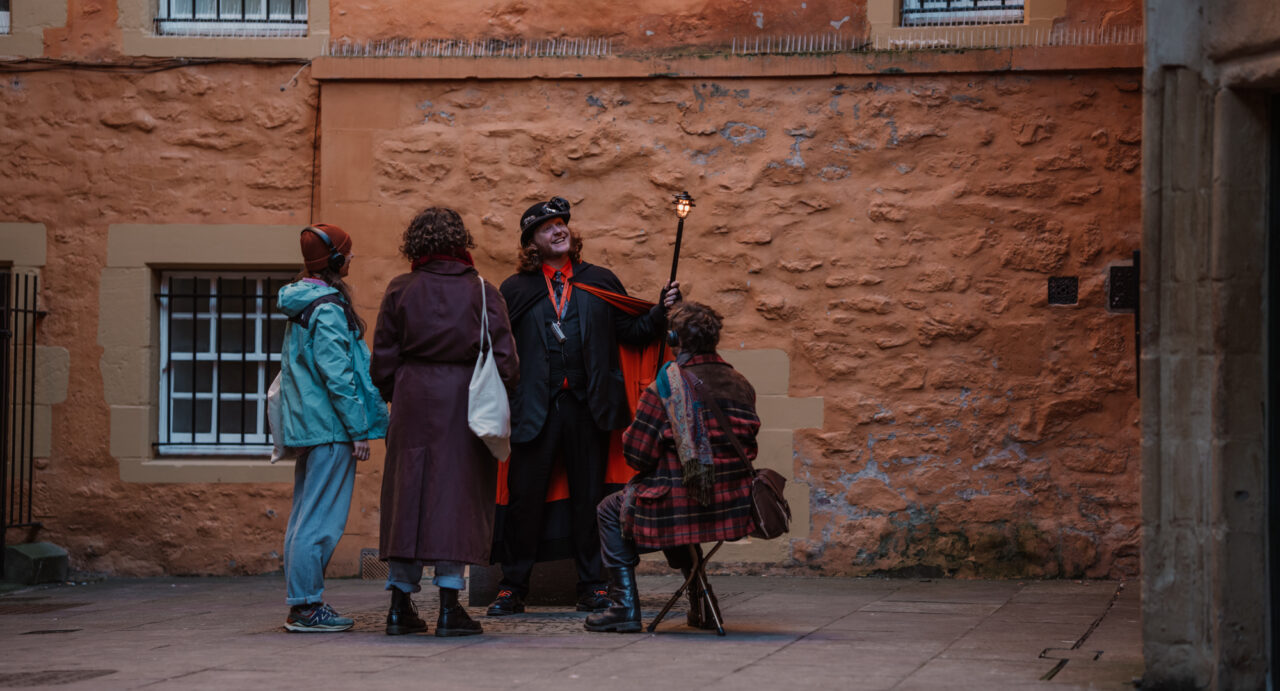A Mercat Tour Guide leading a tour where one of the group is sitting on a stool. All 3 people in the group are wearing headphones.