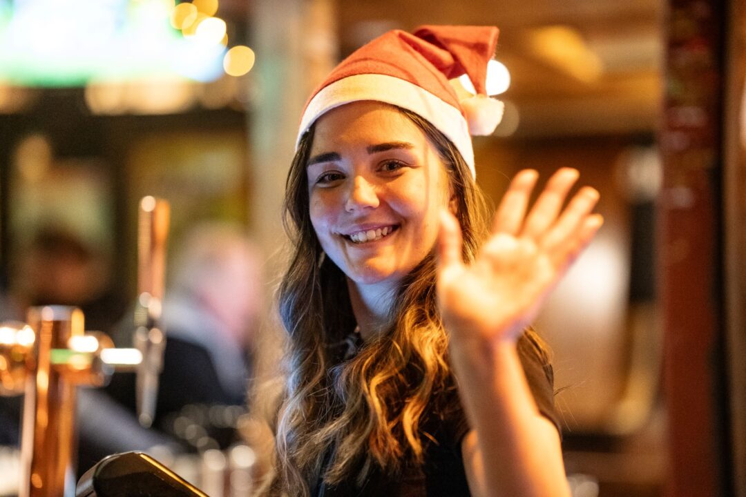 Woman wearing a red and white Santa hat, waving at camera.