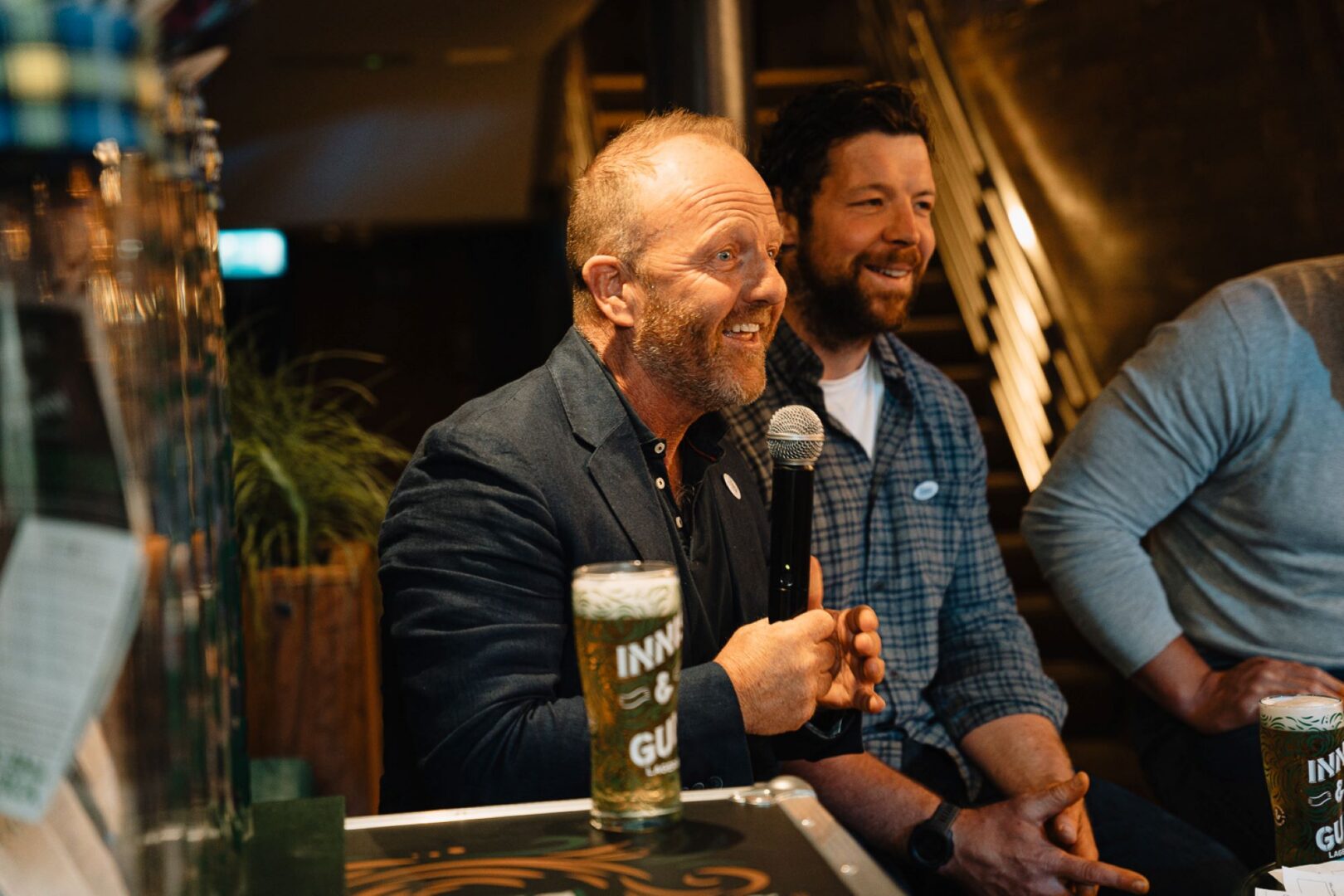 2 men sitting in a bar, one of whom is holding a microphone.