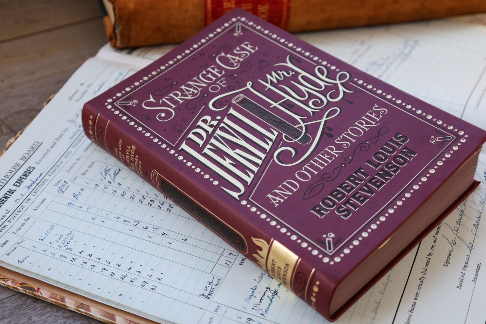 A large hard-booked book with a dark purple cover and gold and black lettering, sitting on a handwritten ledger book.
