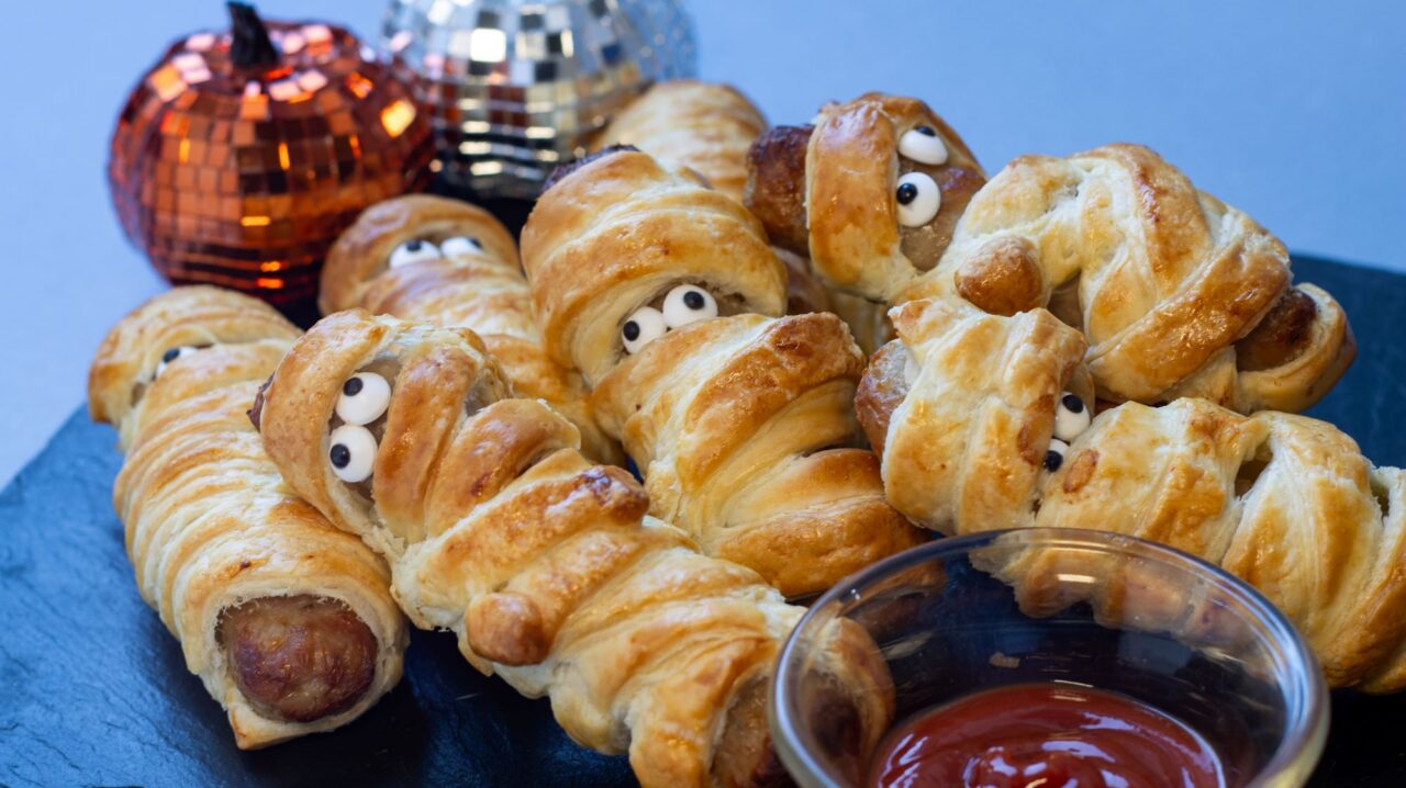 Plate of sausage rolls with eyes, alongside a dish of tomato ketchup and mini pumpkins.