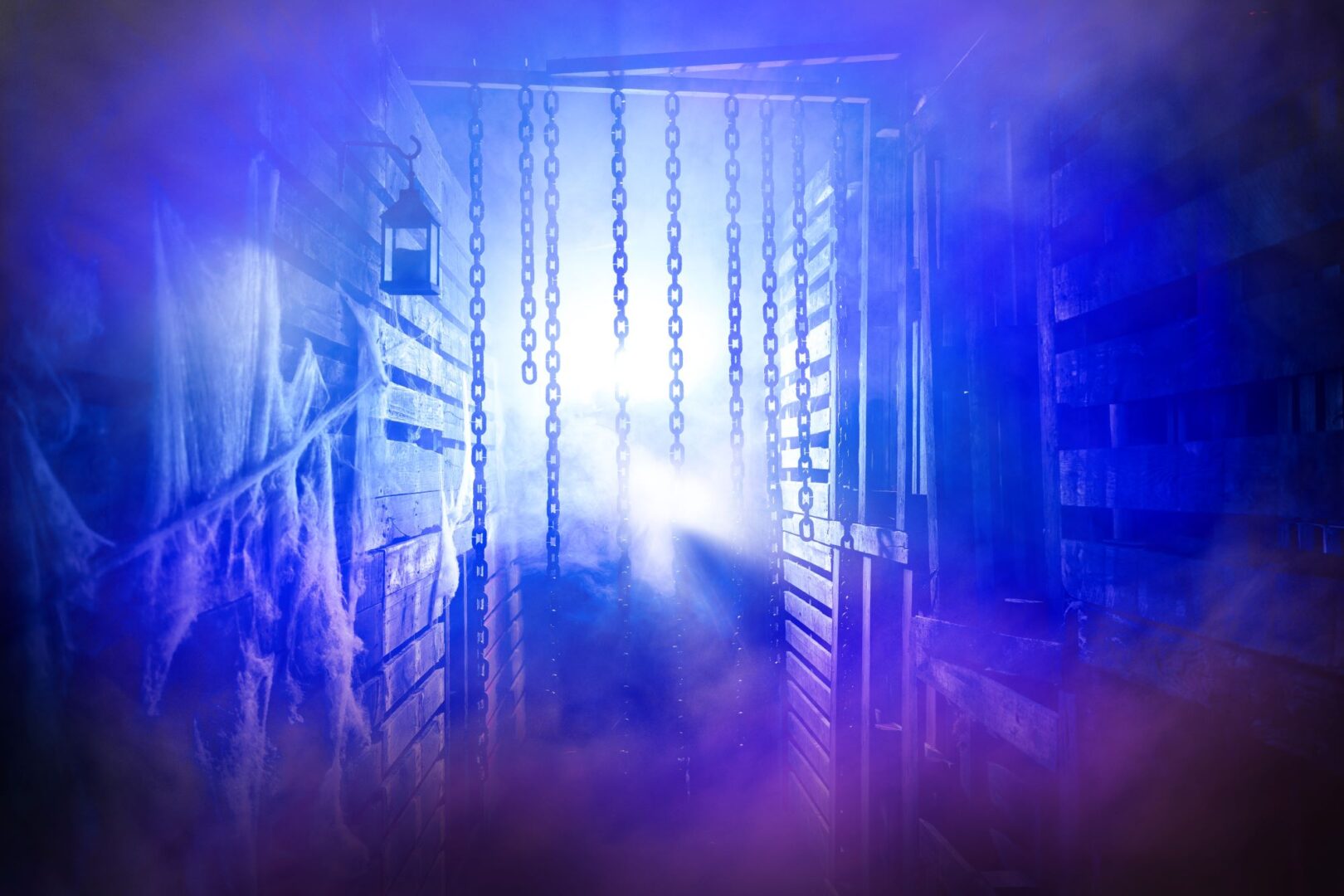 A door ajar, dimly lit, with chains hanging down from the top of the door frame, and blueish smoke coming through.