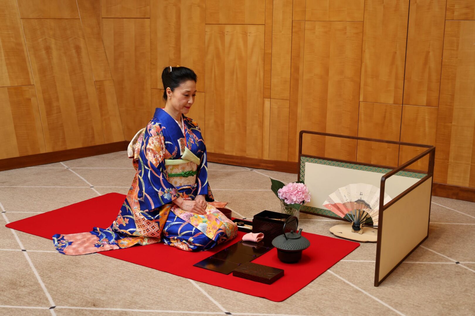 Woman in traditional Japanese dress kneeling on the ground in front of a teapot and vase of flowers.