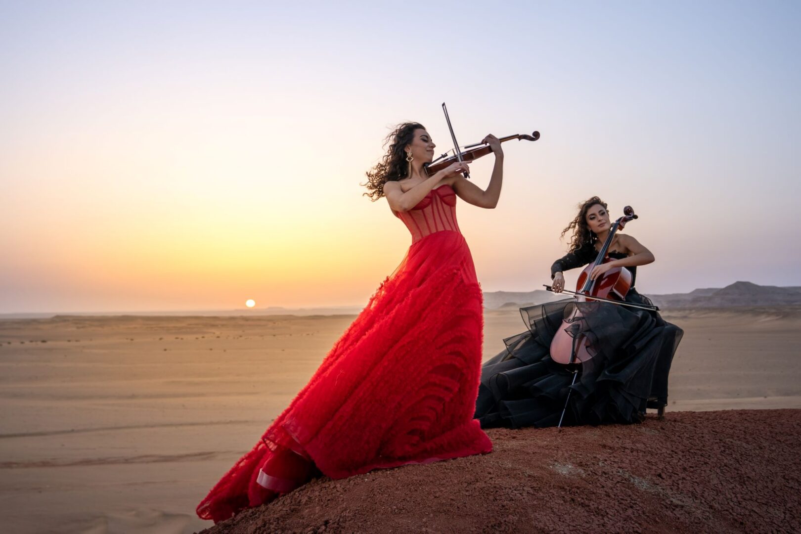 2 women wearing ball gowns playing a violin and a cello on a beach.