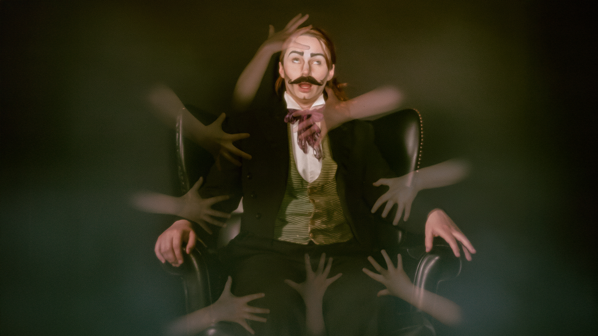 A seated man wearing traditional Victorian dress, with lots of hands on him.