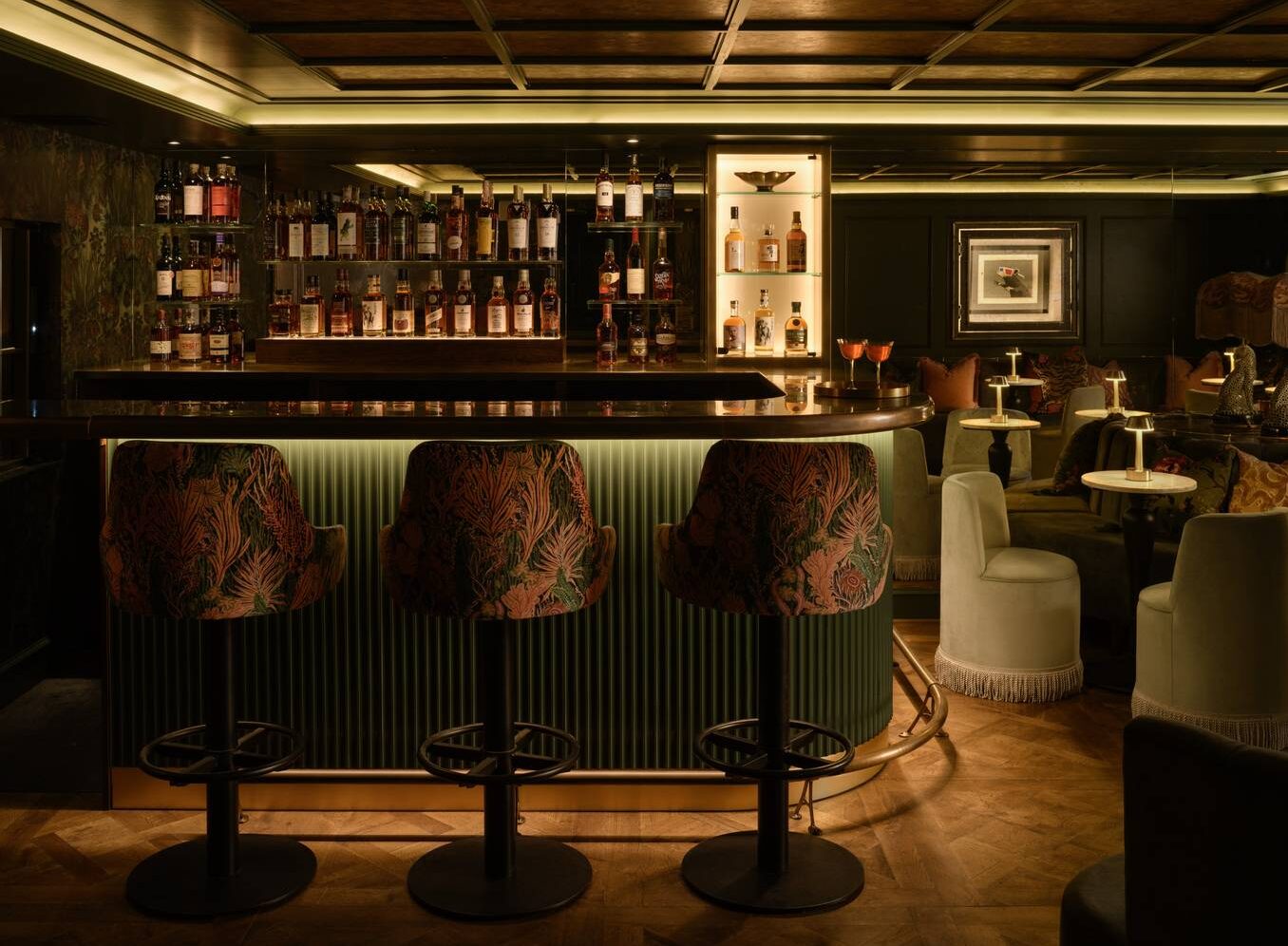 Softly lit bar with backlit shelves of whisky. There are textured upholstered bar stools in rich colours, and velvet seting around white curcular tables with brass lamps. ,© The Burrow - Photography by ZAC and ZAC