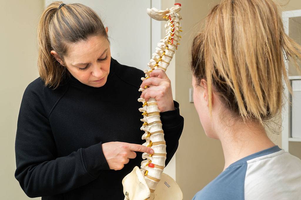 Vicky the chiropractor is educating a patient about what is going on with her back using a model of a spine to demonstrate it.