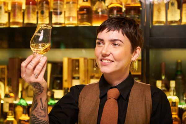 Tour Guide at Scotch Whisky Experience holds up Whisky in a glass with many whisky bottles on shelf behind her.